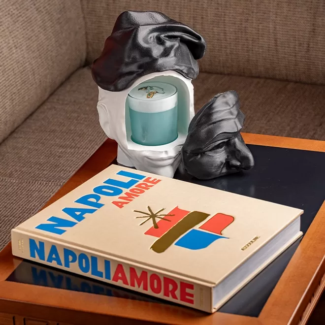 A decorative candle holder shaped like a split head with a black cap holds a blue candle. A matching face piece and the book Napoli Amore rest on a wooden table next to a brown couch.
