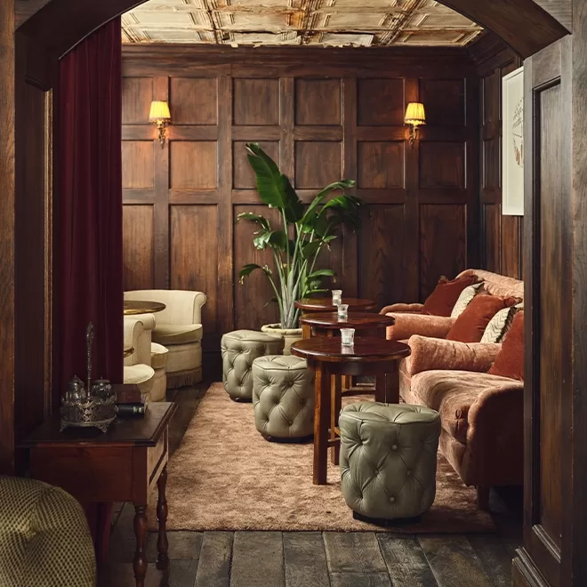 Cozy, wood-paneled lounge with tufted ottomans, velvet sofas, small tables with glassware, armchairs, potted plant, and warm wall sconces, creating an inviting, vintage-inspired atmosphere.