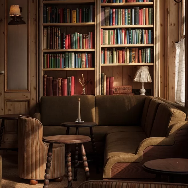 Cozy library corner with a green sectional sofa, round wooden tables, a lit candle, and bookshelves filled with colorful books, warmly lit by natural light from a nearby window.