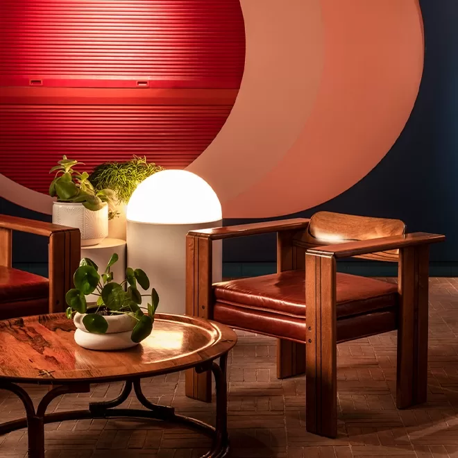 A cozy seating area with two wooden chairs, a round wooden coffee table with potted plants, and a glowing lamp. In the background, there is a large geometric red and white wall design.