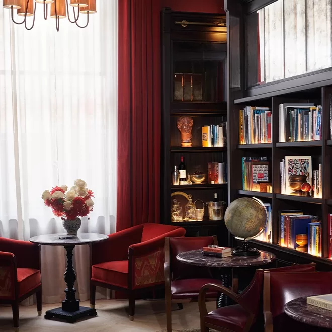 A cozy library room with dark wood shelves filled with books, a globe, and decorative objects. Red armchairs and round tables are arranged near a window with sheer white curtains. A vase of flowers sits on one table.