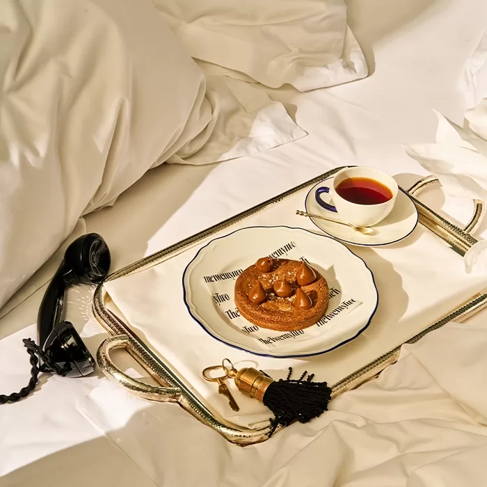 A breakfast tray on a rumpled bed with a pastry on a plate, a cup of tea or coffee, an old-fashioned black telephone, and a pair of keys with a black tassel.