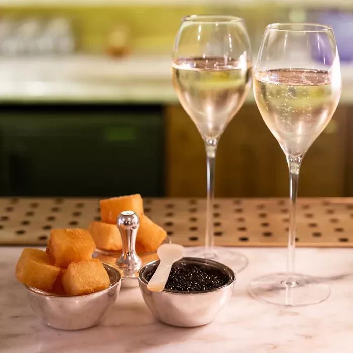 Two glasses of sparkling wine sit on a marble counter next to bowls of golden fried bites and black caviar, with a small spoon and metal caviar server. The background is softly blurred.