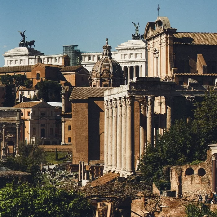 Ancient Roman ruins with tall columns and historic buildings in Rome, Italy, under a clear blue sky. Statues and dome structures are visible in the background.