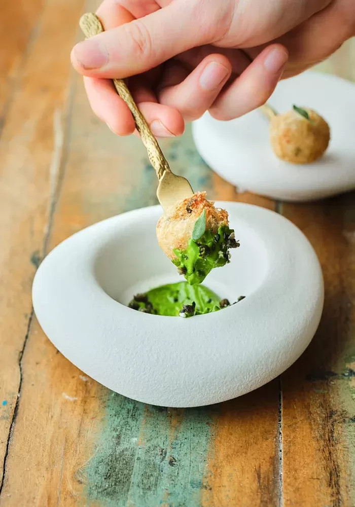 A hand uses a fork to dip a golden-brown, crispy ball garnished with greens into a vibrant green sauce in a modern, white, stone-shaped bowl on a rustic wooden table.