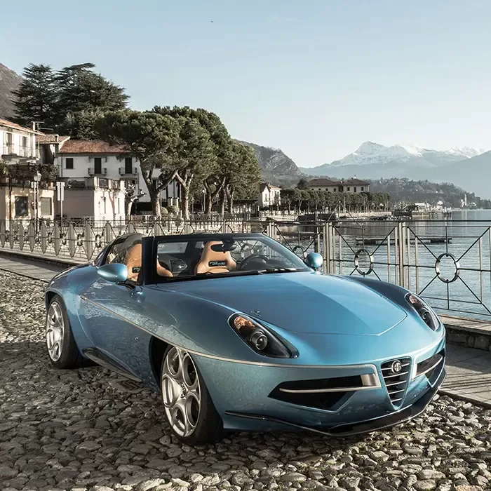 A blue convertible sports car parked on a cobblestone street beside a scenic lake, with mountains and houses in the background under a clear sky.