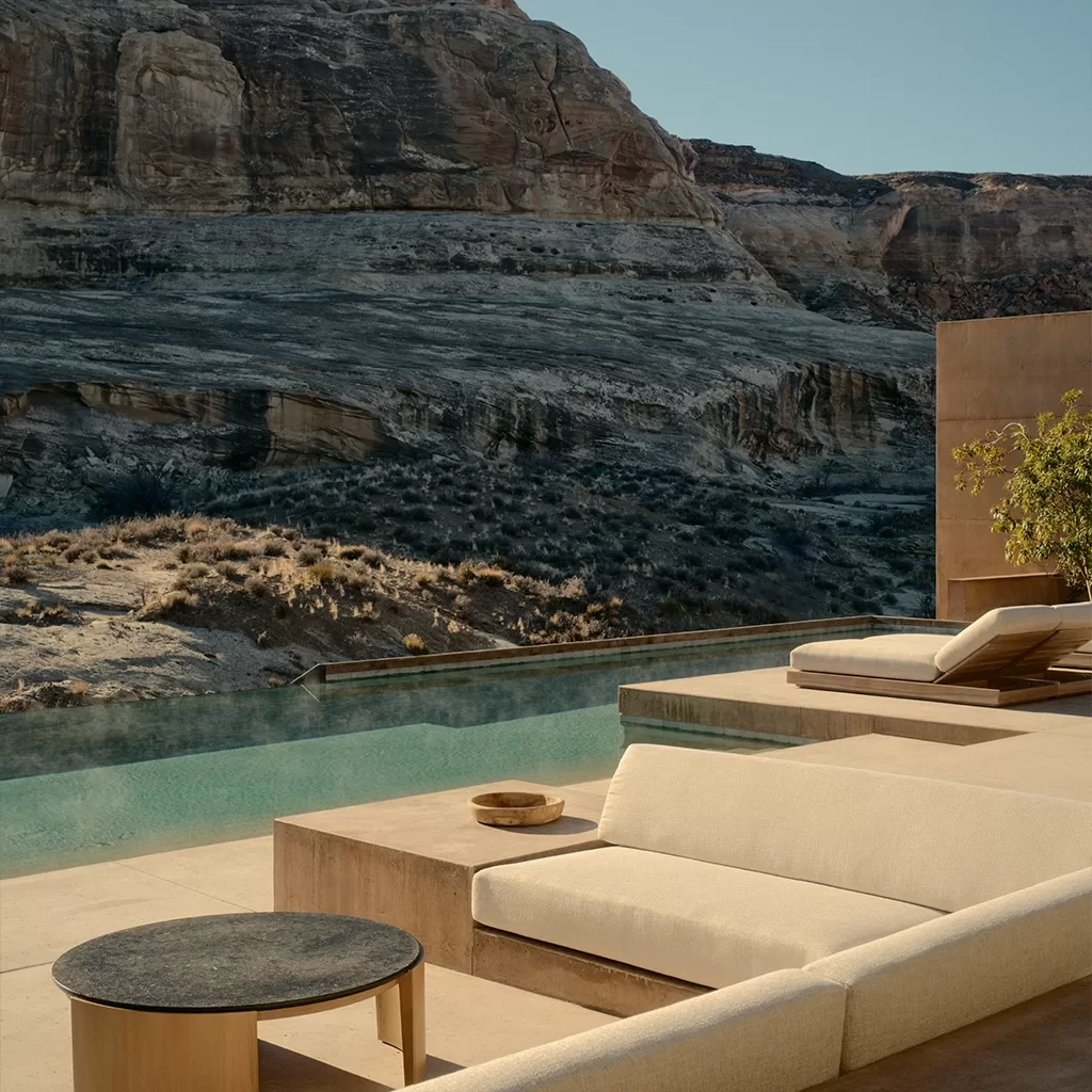 A modern outdoor patio with beige lounge chairs and a small round table overlooks an infinity pool, set against a backdrop of rocky desert cliffs under a clear blue sky.