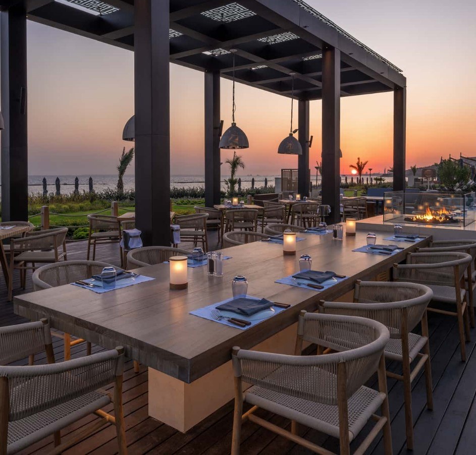 Restaurant with long rectangle table with neutral wicker chairs with sea view.