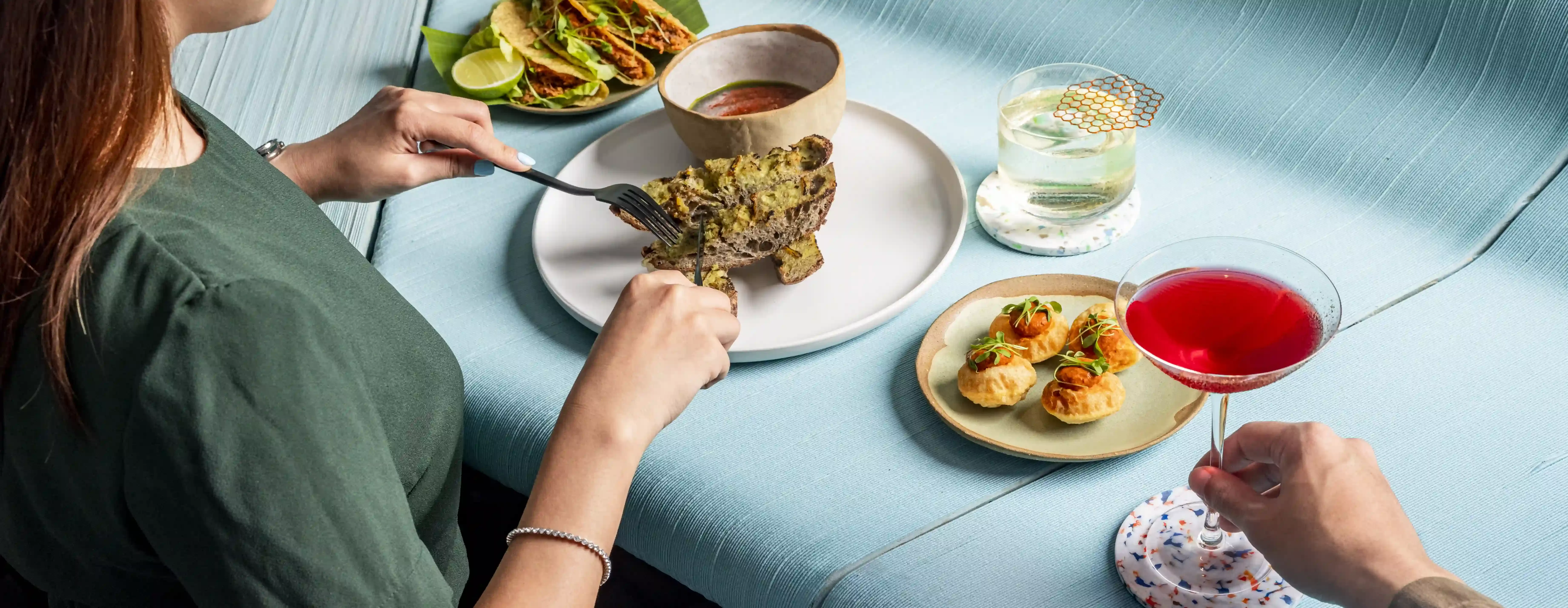 Women eating food of a plate surrounded by various dishes and a cocktail on a sculptural blue table at Analogue Singapore