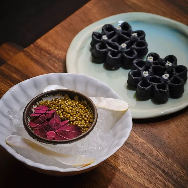 A bowl of green caviar and red leaves sits on ice in a white dish with two white spoons. In the background, black flower-shaped waffles with tiny white flowers are served on a light blue plate.