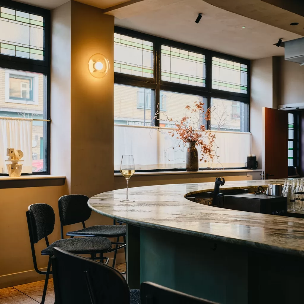A modern bar interior with a curved marble countertop, empty black chairs, a glass of white wine, a vase with dried branches, and large windows letting in natural light.