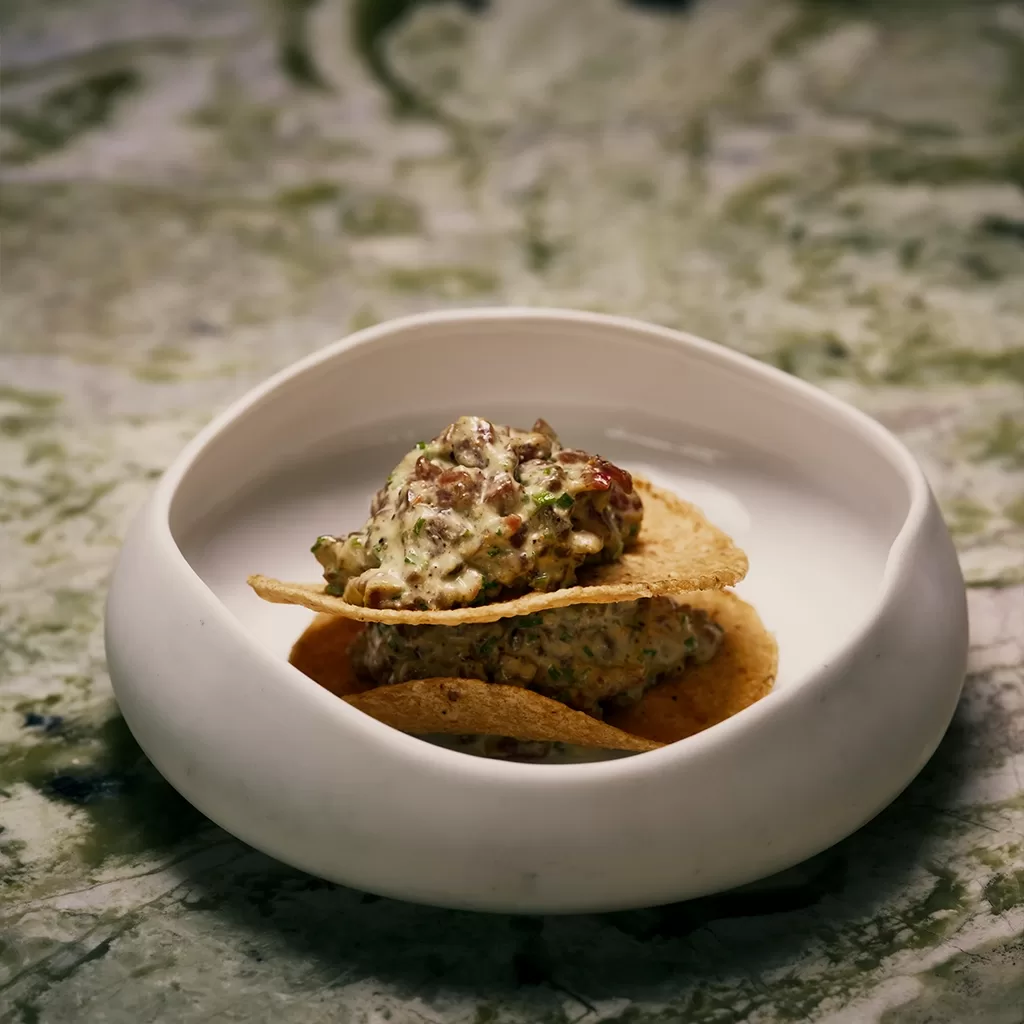 A small white bowl holds two tortilla chips topped with a creamy, chunky dip, set on a green and white marbled surface.