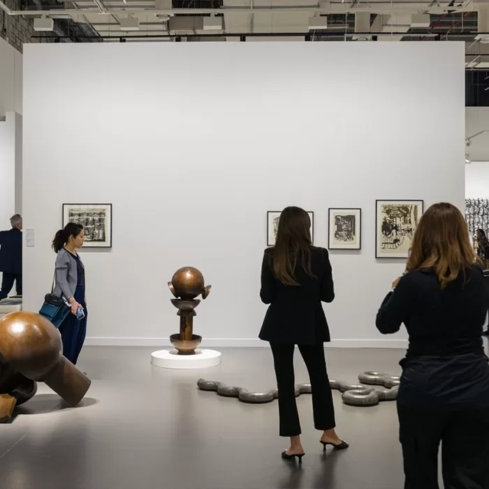 People view sculptures and framed art in a modern gallery. Three women stand near large metal sculptures and black-and-white framed artworks on a white wall. The gallery space is minimal and well-lit.