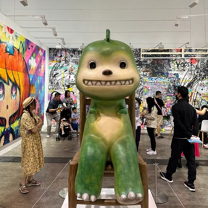 A large green and yellow cartoonish creature sculpture with a big smile is displayed in an art gallery, surrounded by visitors and colorful graffiti-style wall art.