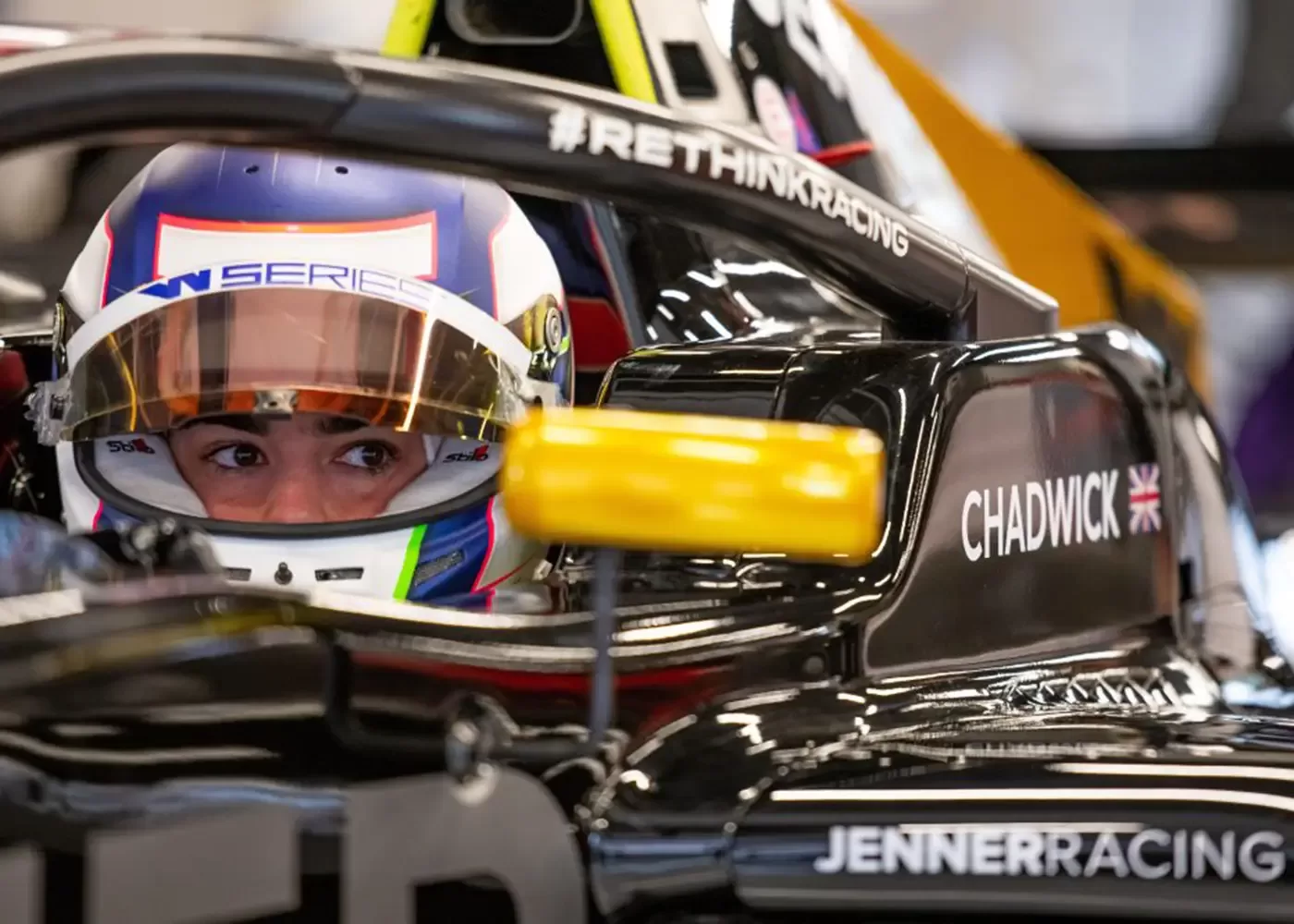 A race car driver wearing a helmet sits in a black Formula car with CHADWICK and a British flag on the side, and JENNER RACING written below. The driver looks focused, gripping the steering wheel.