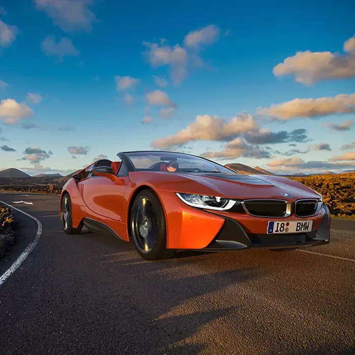 A bright orange BMW i8 convertible sports car is driving on a curvy road with a scenic landscape, including mountains and a blue sky with scattered clouds, in the background.