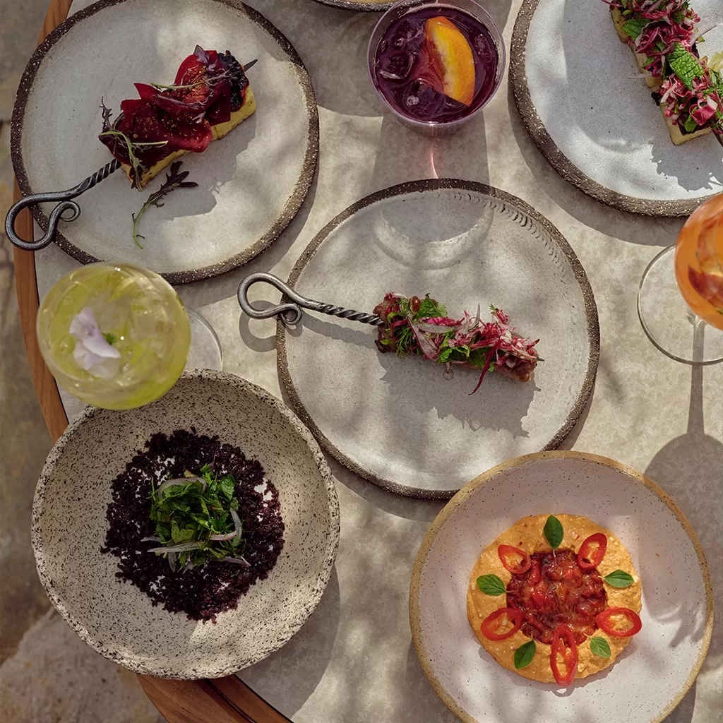An overhead shot of a table laid with various small plates and drinks. Dishes include skewers of meat and vegetables, a bowl of dark granular food with herbs, and a flatbread topped with red ingredients and basil.