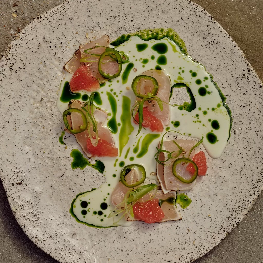 Thinly sliced raw fish with grapefruit, jalapeño rings, and a vibrant green sauce drizzled over a creamy sauce on a speckled plate.