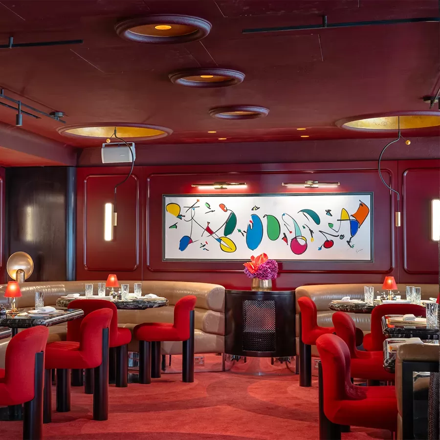 A stylish restaurant with red chairs, set tables, curved beige booth seating, modern lighting, and a colorful abstract painting on a deep red wall. Pink flowers decorate a table in the center.
