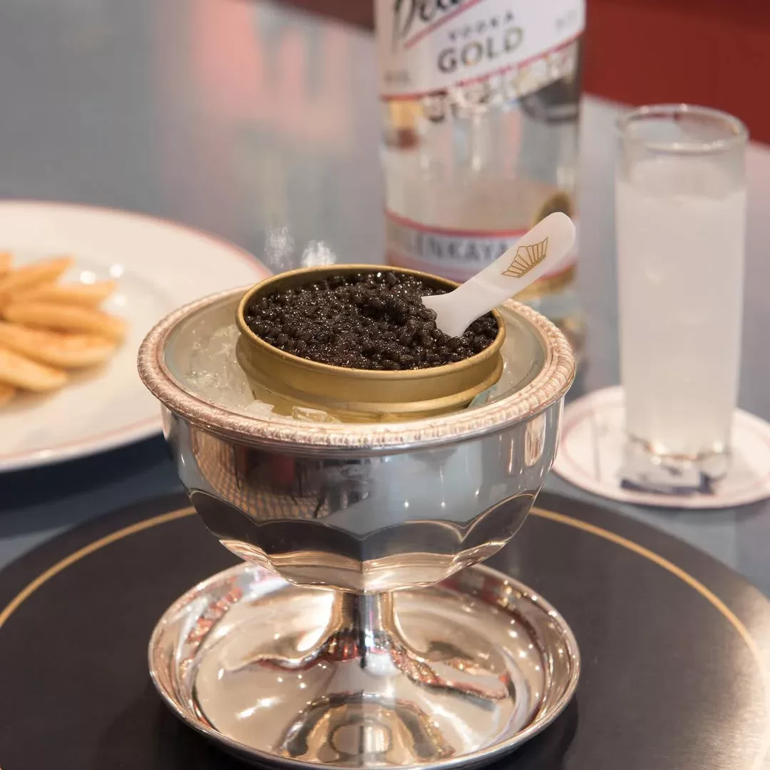 A tin of black caviar with a small white spoon sits on ice in a silver dish. In the background, there’s a bottle of vodka, a glass with clear liquid, and a plate of fries.