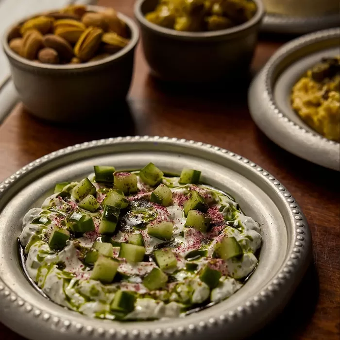 A plate of creamy yogurt dip topped with chopped cucumber, herbs, and spices sits on a wooden table, with bowls of nuts and other dips in the background.