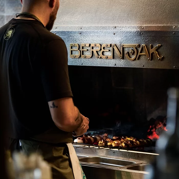 A person grills skewers of meat over an open flame in a kitchen. The metallic wall behind features the restaurant name BERENJAK in raised letters.