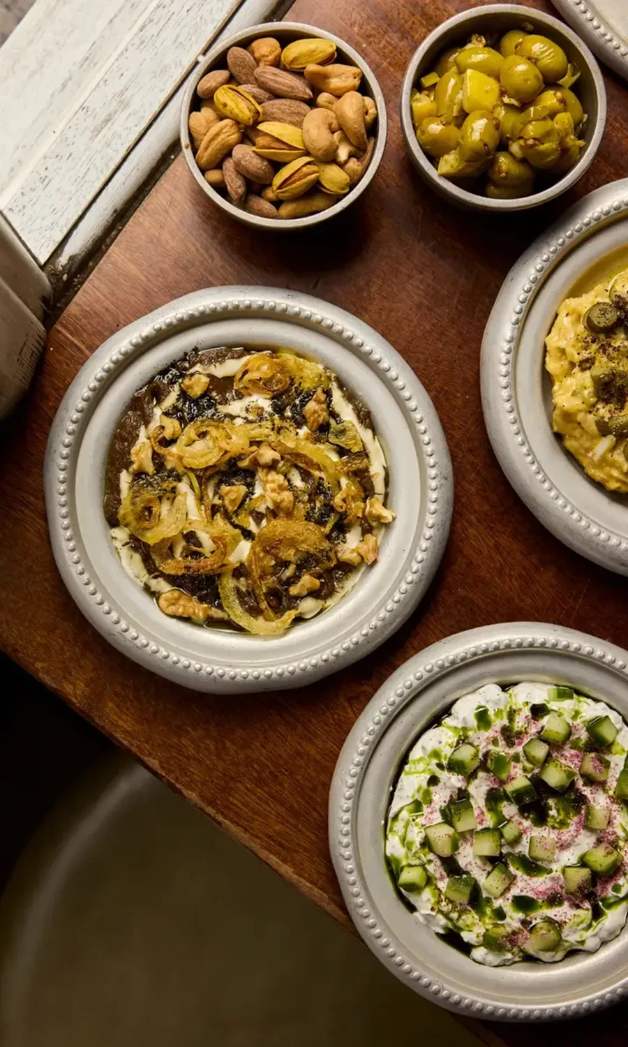 Three small bowls of dips topped with various ingredients sit on a wooden surface, accompanied by side bowls of mixed nuts and green olives. The dips appear creamy and are garnished with vegetables and spices.