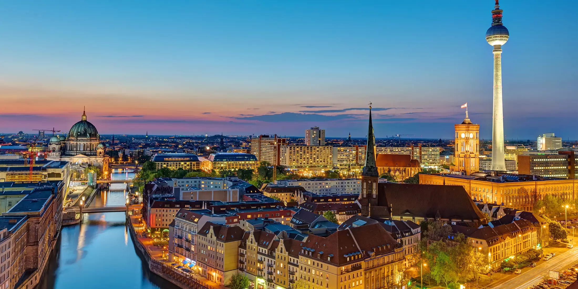 Aerial view of Berlin at dusk, featuring the illuminated cityscape with the TV Tower, historic buildings, and the Spree River reflecting the evening lights under a colorful sky.