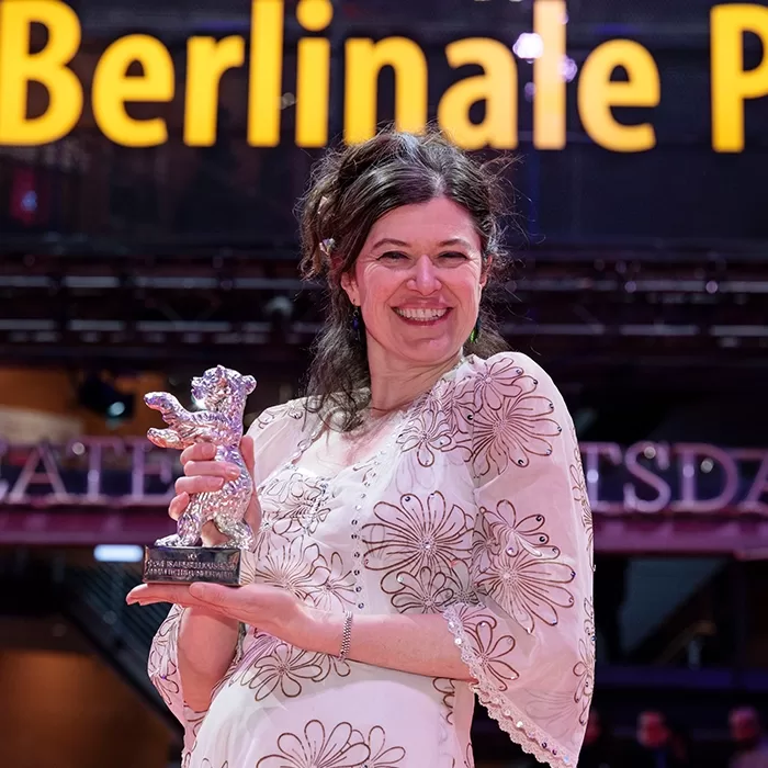 A smiling woman in a white dress with floral patterns holds a silver bear-shaped trophy. The background features large, illuminated yellow letters spelling Berlinale.