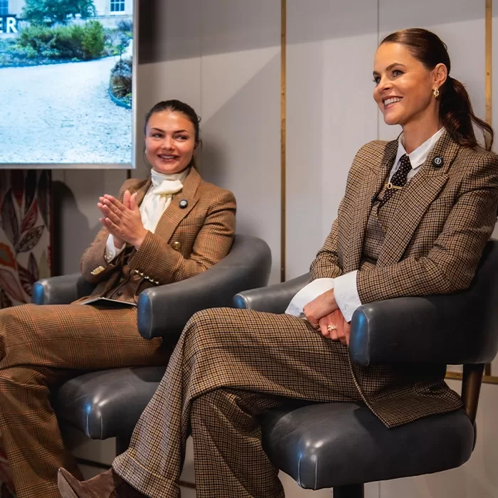 Two women wearing brown plaid suits sit on chairs indoors, smiling and engaged in conversation. One woman claps her hands while the other rests her hands on her lap. A screen and part of a window are visible in the background.