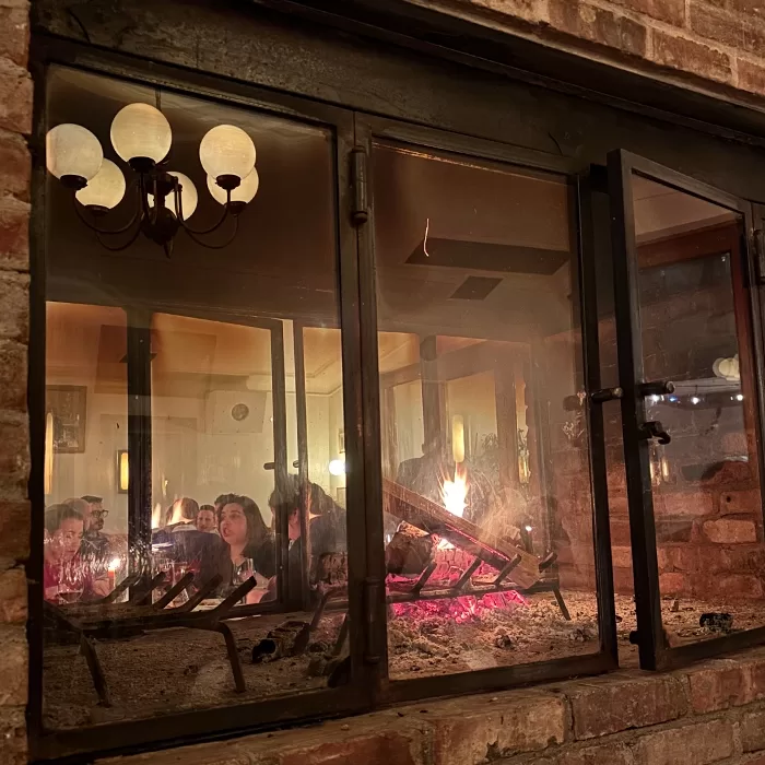 People are seated at a dining table, seen through a glass fireplace with burning logs and glowing embers, inside a warmly lit brick-walled room with a chandelier reflected in the glass.