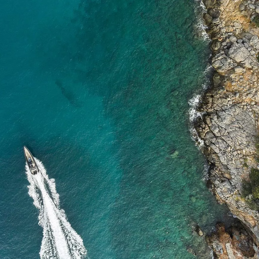 Birdseye view of a boat driving through a blue sea next to rocks