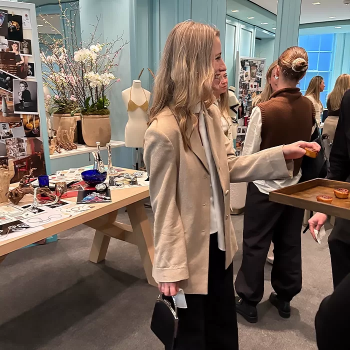 A woman in a beige blazer holds a small black bag and takes an appetizer from a tray at an indoor event; tables with decorative items and photo boards are in the background.
