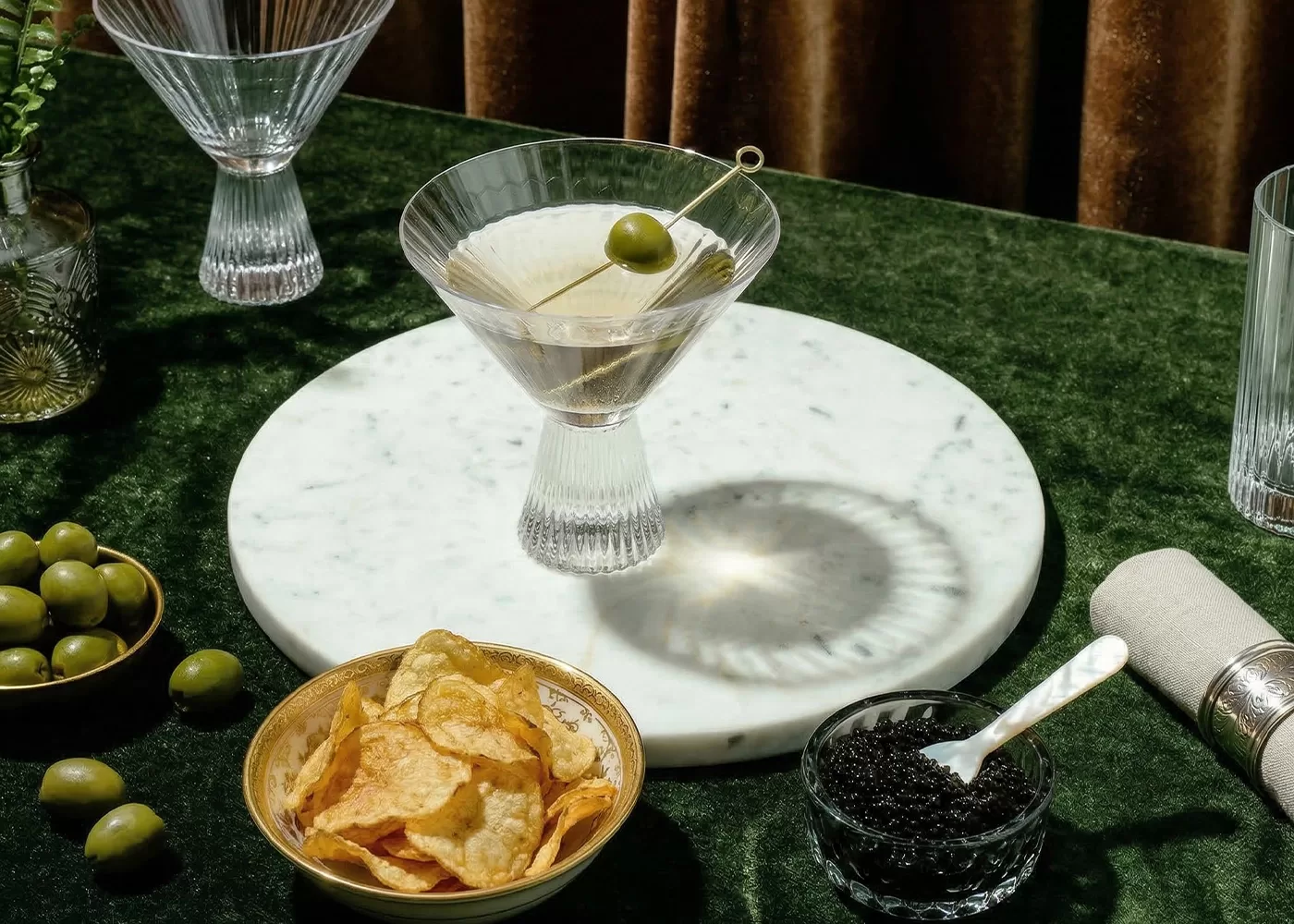 A martini with an olive sits on a marble tray, surrounded by a bowl of potato chips, a dish of caviar with a spoon, extra olives, and empty glasses on a green velvet tablecloth.