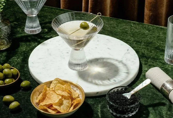 A martini with a green olive sits on a round marble board atop a dark green tablecloth, next to a bowl of potato chips, a small bowl of caviar with a spoon, and a few loose olives.