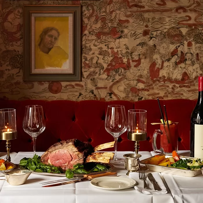 A table set for two features a large prime rib roast with greens, candles, wine glasses, and a bottle of red wine on a white tablecloth. The background shows a red velvet booth and ornate wallpaper with a framed portrait.