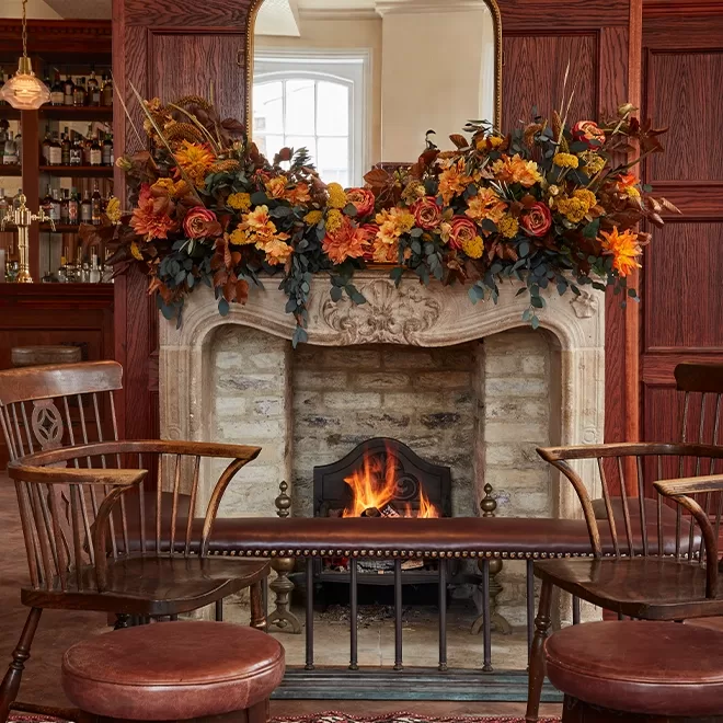 A cozy stone fireplace with a fire burning, decorated with a large autumn floral arrangement. Wooden chairs and stools are arranged around the fireplace in a warmly lit, wood-paneled room.
