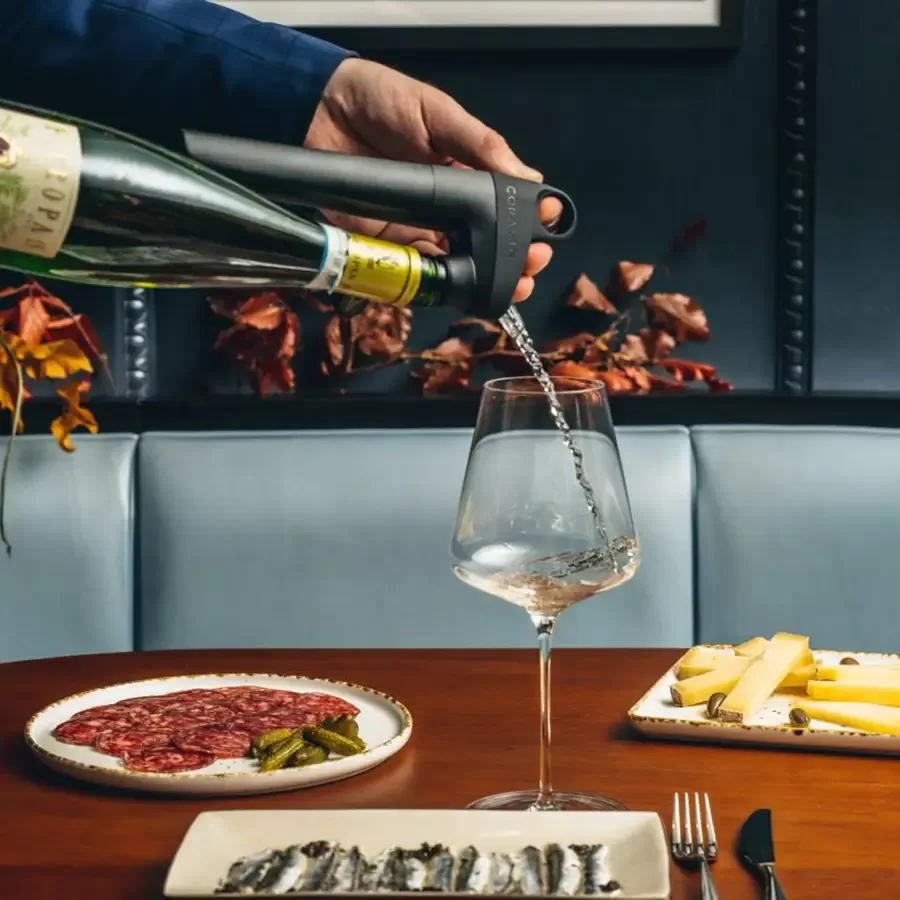 A hand pours white wine from a bottle into a glass at a restaurant table set with charcuterie, cheese, pickles, and small fish dishes.
