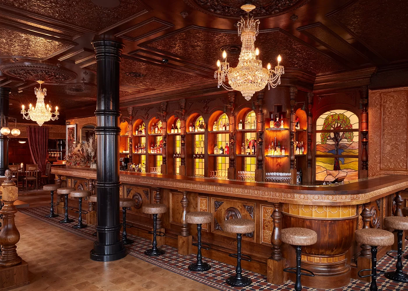 A lavish bar with ornate chandeliers, stained glass windows, arched shelves filled with bottles, a polished wooden counter, and several cushioned barstools in a richly decorated, vintage-style interior.