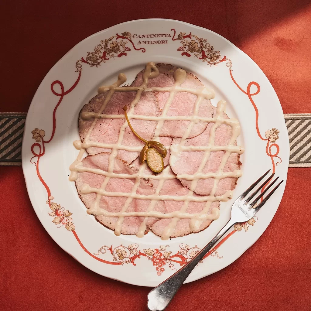 A plate of thinly sliced roast veal topped with a crisscross drizzle of creamy sauce and a caper, presented on a decorative white plate with a fork, set on a red tablecloth.