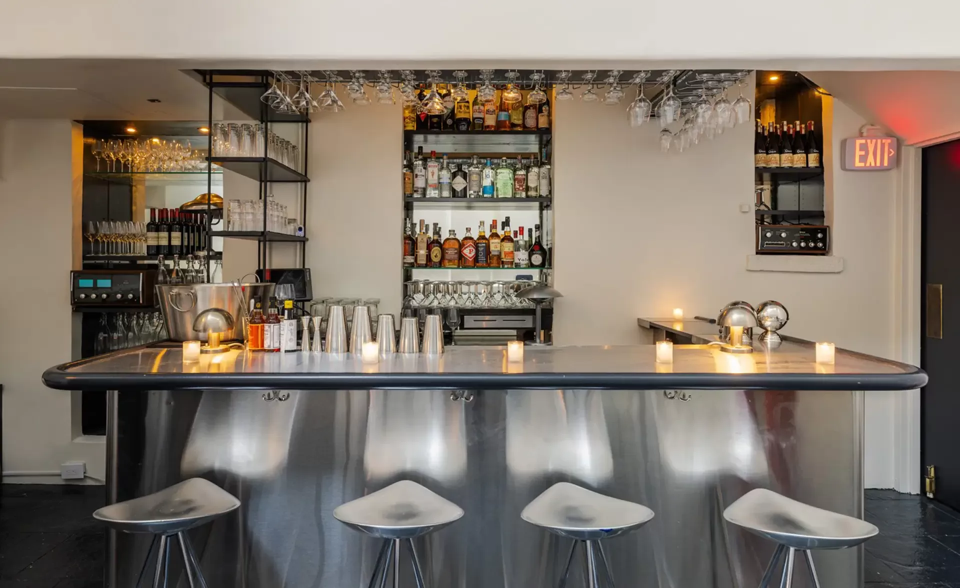 A sleek, modern bar with a shiny metallic counter and stools. Shelves behind display various bottles and glassware. Soft lighting and candles create a warm, inviting atmosphere. An exit sign is visible on the right.