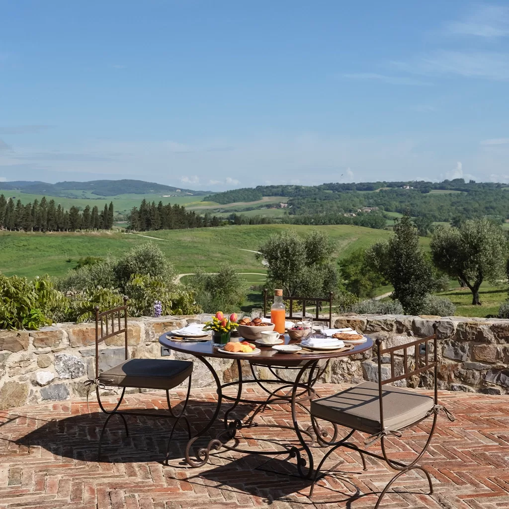 A round table set for breakfast with four chairs sits on a brick patio overlooking rolling green hills, trees, and a blue sky in a scenic countryside setting.