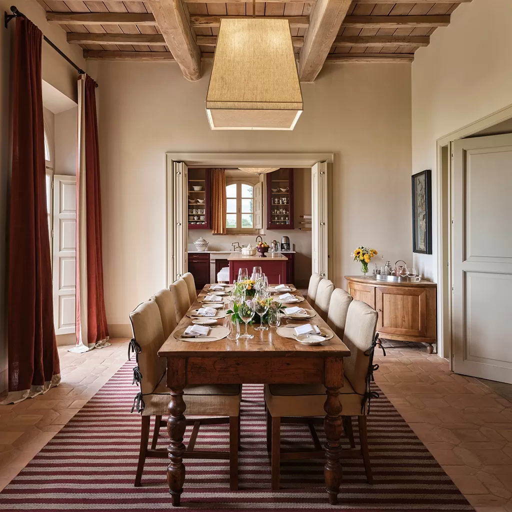 Elegant dining room with a wooden table set for eight, beige cushioned chairs, striped rug, large pendant light, rustic ceiling beams, and open double doors leading to a kitchen with sunflowers on a sideboard.