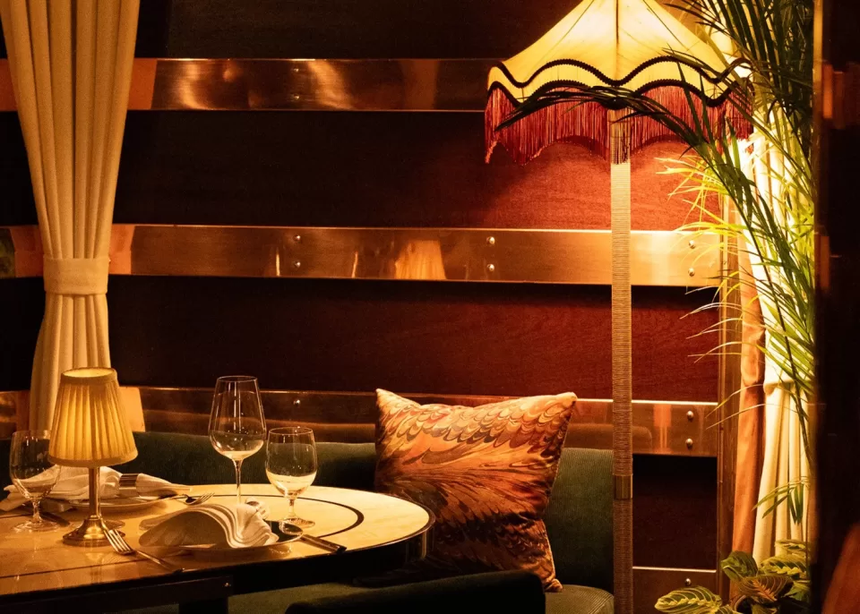 A cozy restaurant corner with a round table set for two, wine glasses, a patterned pillow on a green bench, and a vintage lamp casting warm, ambient light in a softly lit room with dark wood paneling.