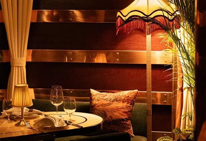 A cozy restaurant table set for two with wine glasses and a small lamp. A patterned pillow rests on a green bench under a vintage floor lamp, with warm, ambient lighting and elegant curtains in the background.