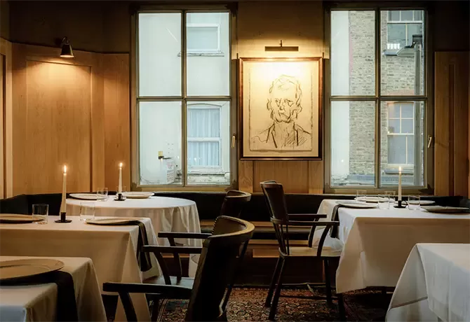 A cosy restaurant interior features tables with white tablecloths and lit candles. Soft lighting from a window illuminates a monochrome portrait on the wall, creating an intimate ambiance at Cycene, Shoreditch.