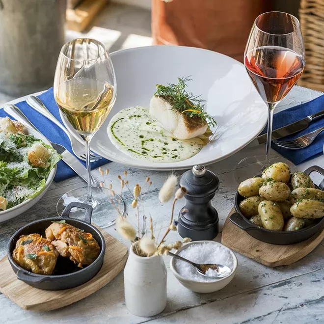 A dining table set with a seafood dish in a white bowl, two glasses of wine, a Caesar salad, roasted potatoes, a small skillet with vegetables, a pepper grinder, and a vase with dried flowers on a marble surface at Chalk Restaurant, Wiston Estate.