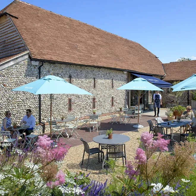 Outdoor cafe with umbrellas and tables, set against a historic stone building with a red-tiled roof. People sit and enjoy the sunny day. Pink and purple flowers in the foreground enhance the scene at Chalk Restaurant, Wiston Estate.