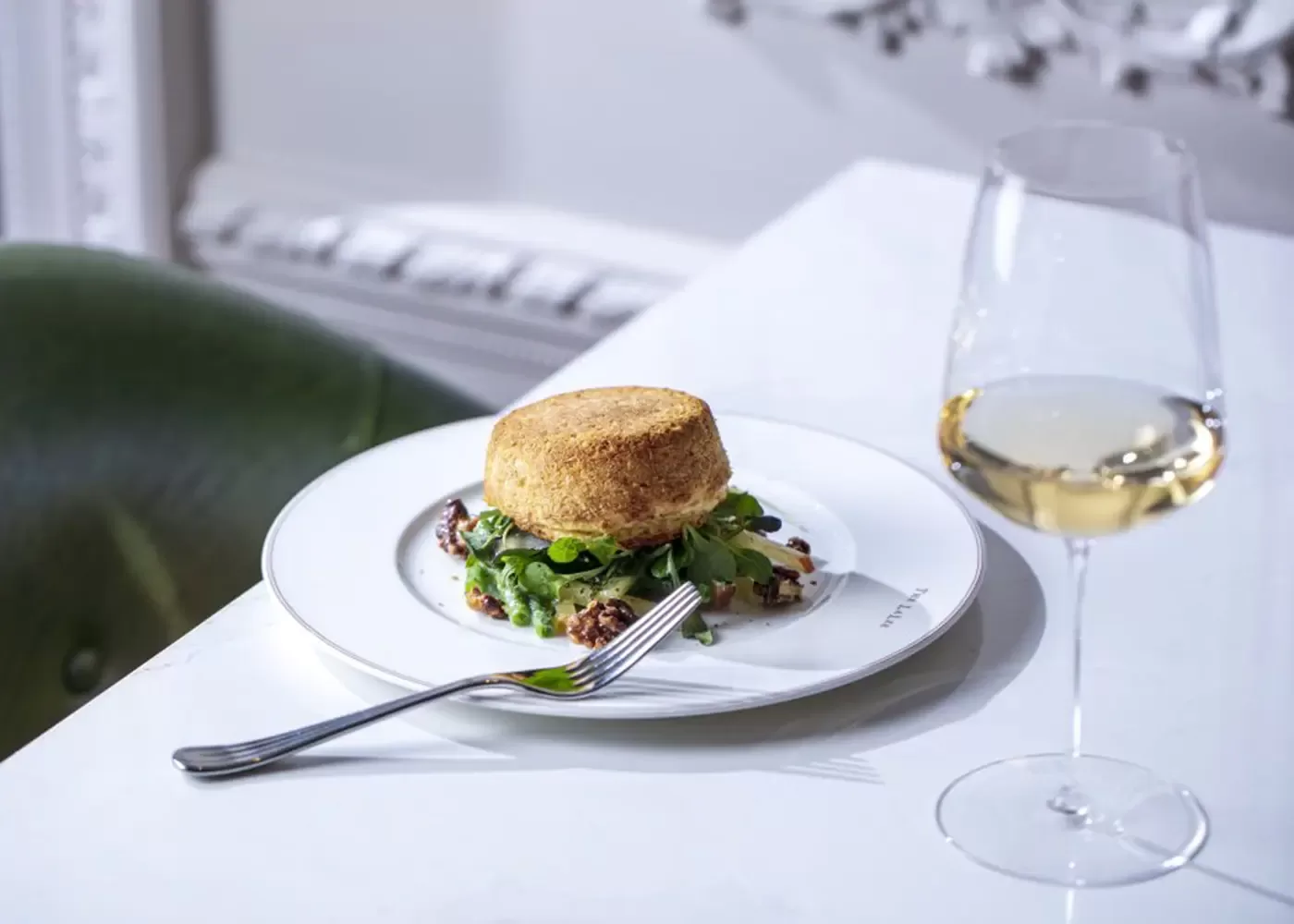 A golden-brown breaded patty sits on a bed of greens and sauce on a white plate with a fork, next to a glass of white wine on a white table.