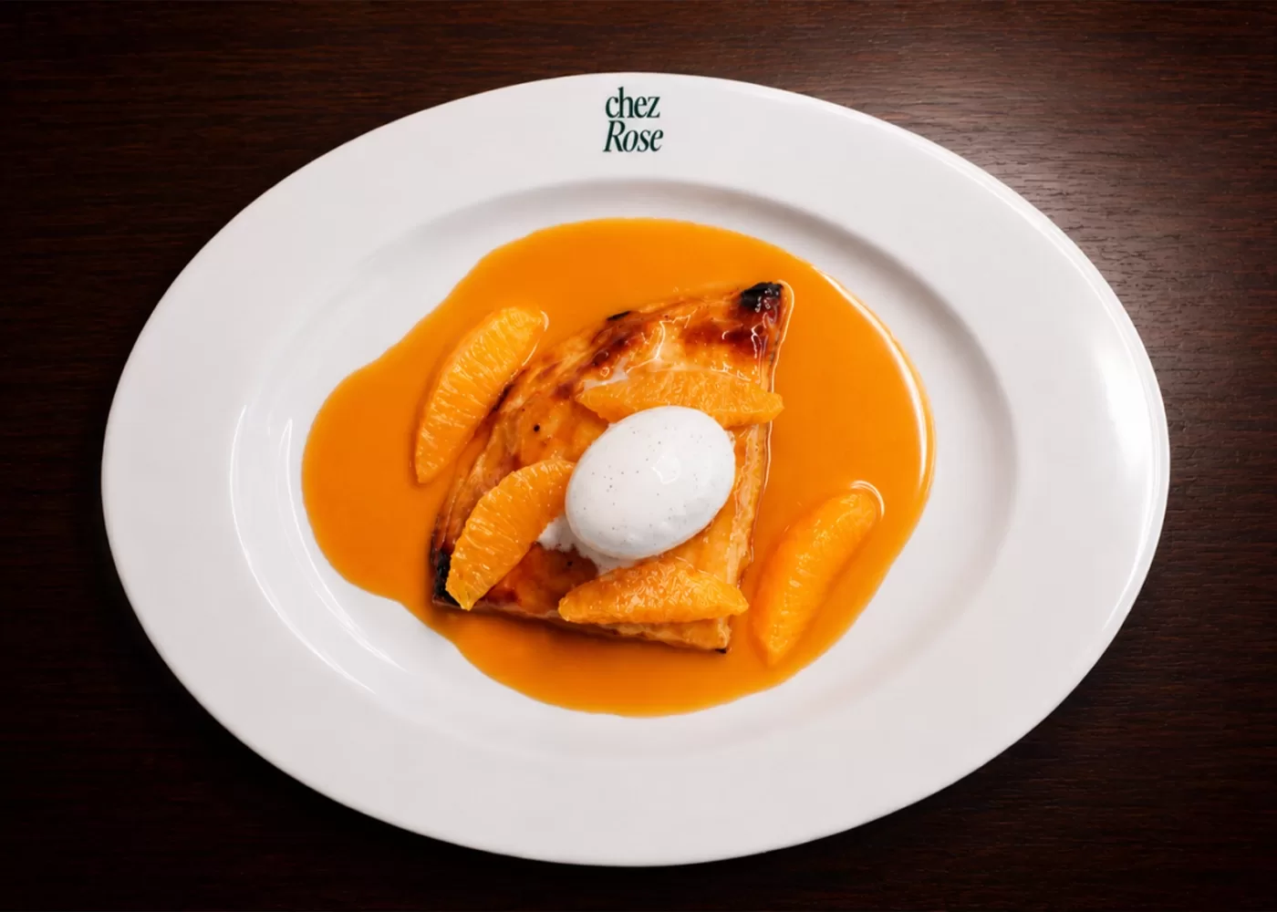 A white plate with a slice of dessert topped with a scoop of ice cream, surrounded by orange sauce and mandarin slices, on a dark wooden table. The plate is labeled “chez Rose” at the top.